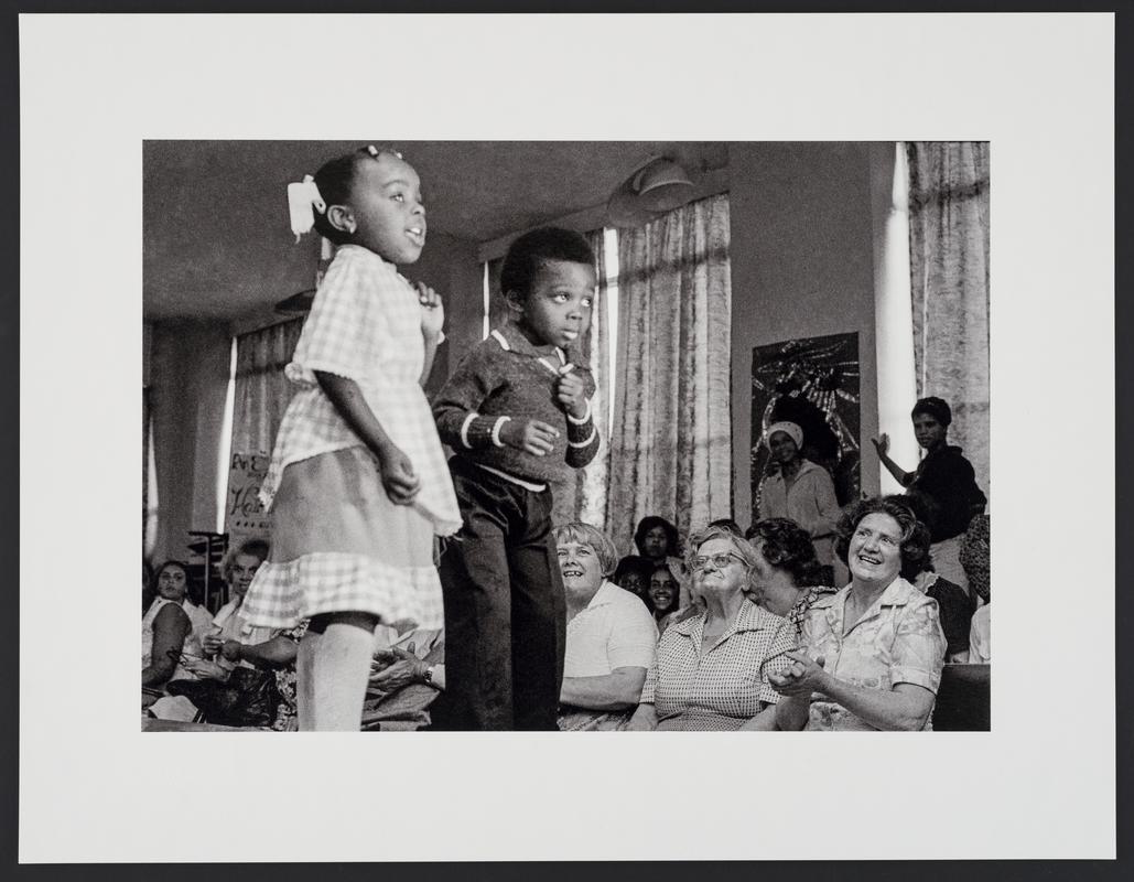 Young children on the catwalk pose for a fashion show in Butetown Community Centre, while the audience applaud. 1979