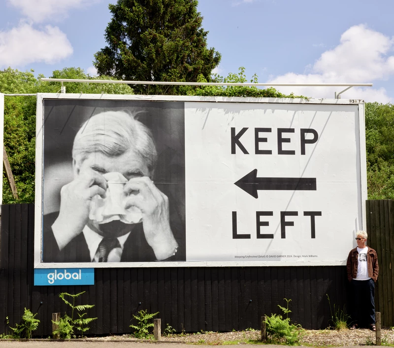 Bwrdd poster o ffotograff du a gwyn o Aneurin Bevan a 'Keep Left' mewn llythrennau bras du gyda saeth du yn pwyntio i'r chwith. Safai'r artist i'r dde o'r bwrdd poster.